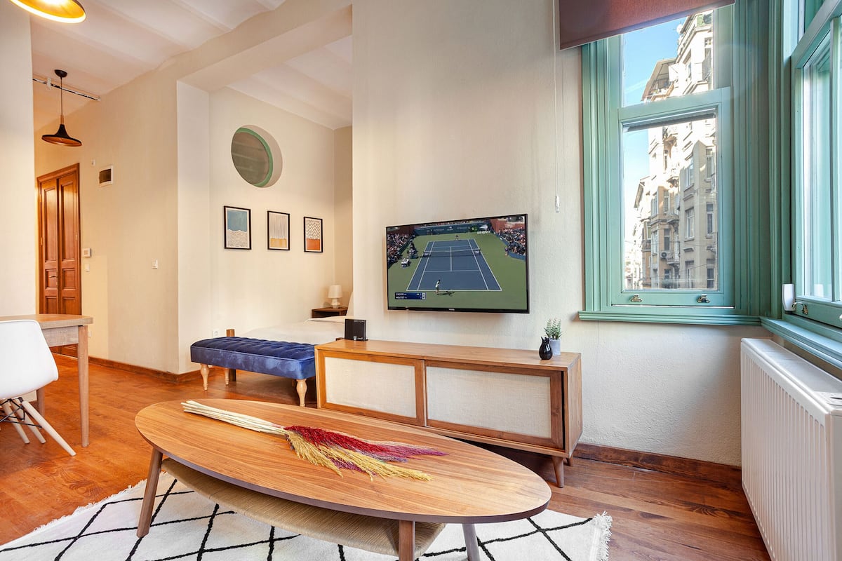 A room with a wooden table, a blue bench, a TV on the wall, and a window with a view of buildings.