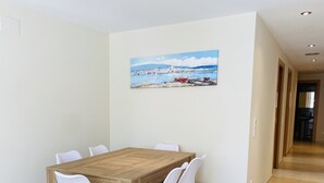 Uma sala de jantar com uma mesa de madeira e cadeiras brancas, um quadro de barcos na parede e piso de azulejos.