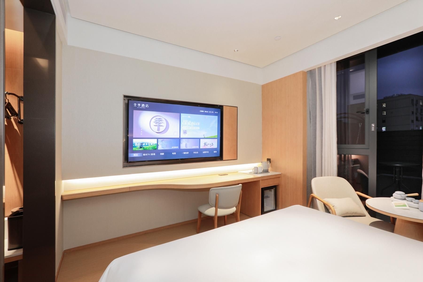 A hotel room with a bed, a desk with a chair, a television mounted on the wall, and a window with a view of the city.