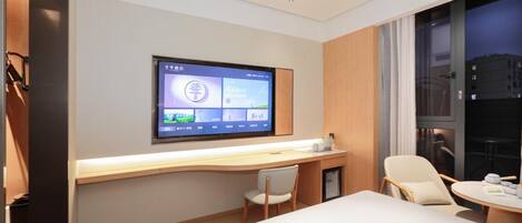 A hotel room with a bed, a desk with a chair, a television mounted on the wall, and a window with a view of the city.