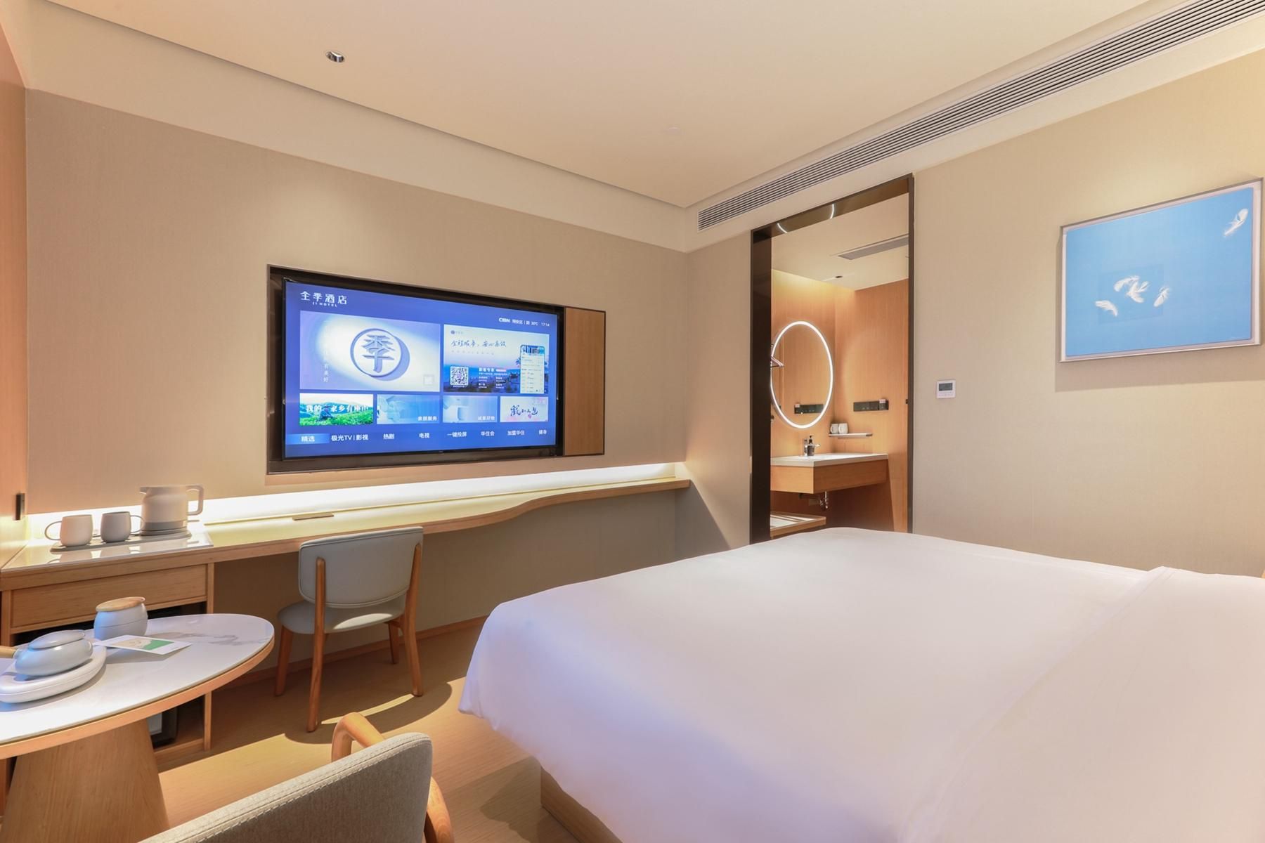 A hotel room with a bed, a desk with a computer monitor, a chair, and a small table with cups and a kettle.