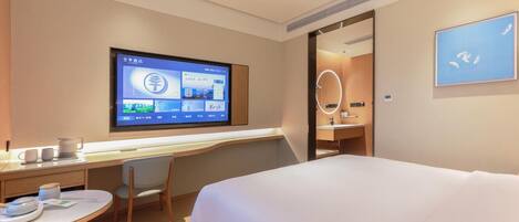 A hotel room with a bed, a desk with a computer monitor, a chair, and a small table with cups and a kettle.
