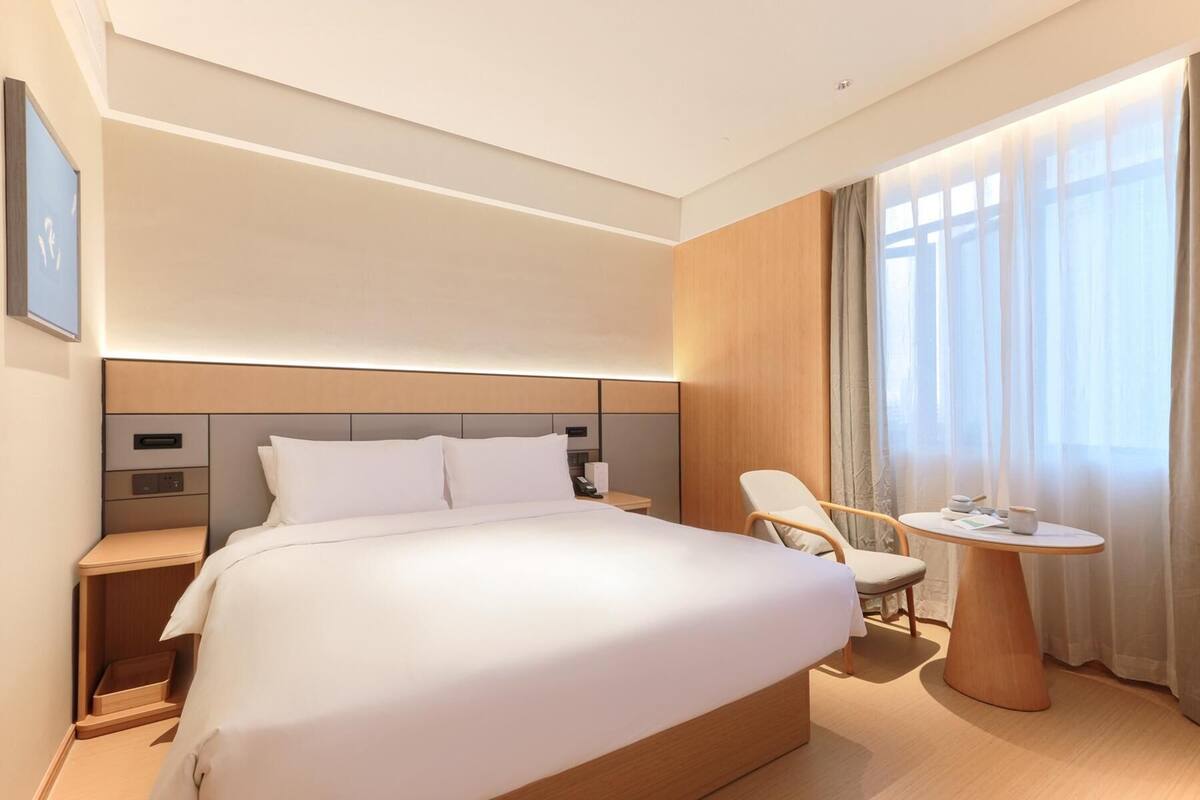 Una habitación de hotel moderna con una cama grande, mesitas de noche, una silla, una pequeña mesa redonda y una ventana con cortinas.