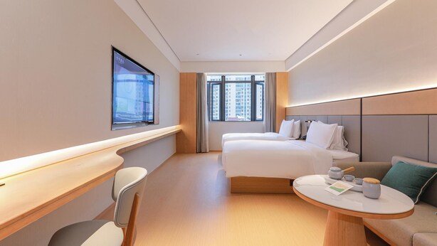 Een hotelkamer met een bed, een bureau, een stoel, een ronde tafel met een theepot, en een raam met uitzicht op de stad.