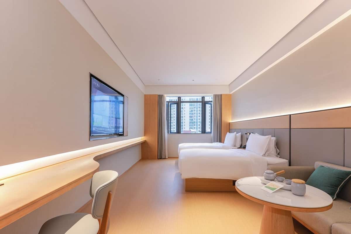 Ein Hotelzimmer mit einem Bett, einem Schreibtisch, einem Stuhl, einem runden Tisch mit Teekanne und einem Fenster mit Blick auf die Stadt.