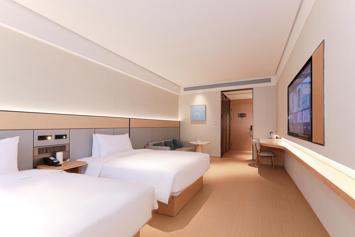 Ein Hotelzimmer mit zwei Betten, einem Schreibtisch, einem Stuhl, einem kleinen Tisch, einem Telefon und einem großen Fernsehbildschirm.