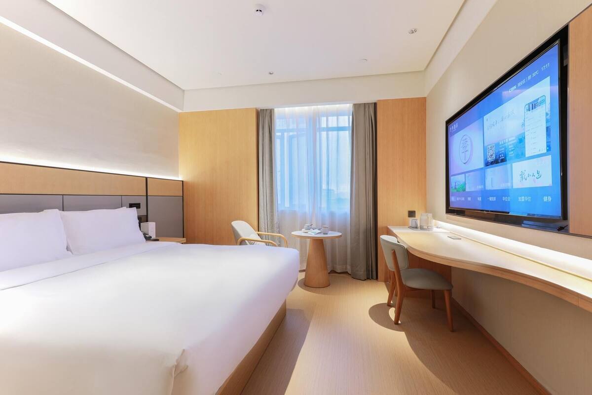 Una habitación de hotel moderna con una cama grande, un escritorio y un televisor mostrando una interfaz digital.