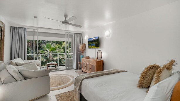 A modern bedroom with a large bed, a sofa, a dining table, and a view of a mountain through a sliding glass door. - Belle Escapes Drift Beachfront Resort Suite 3409 (Palm Cove)