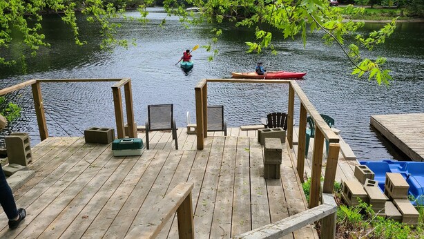 Un muelle de madera con sillas y una mesa, con vista a un río tranquilo y kayaks.