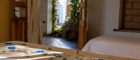A room with a bed, a wooden table with painted flowers, and a view of a courtyard through an open door.
