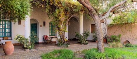 Un cortile con un albero, piante in vaso e un edificio rustico con archi e porte verdi.
