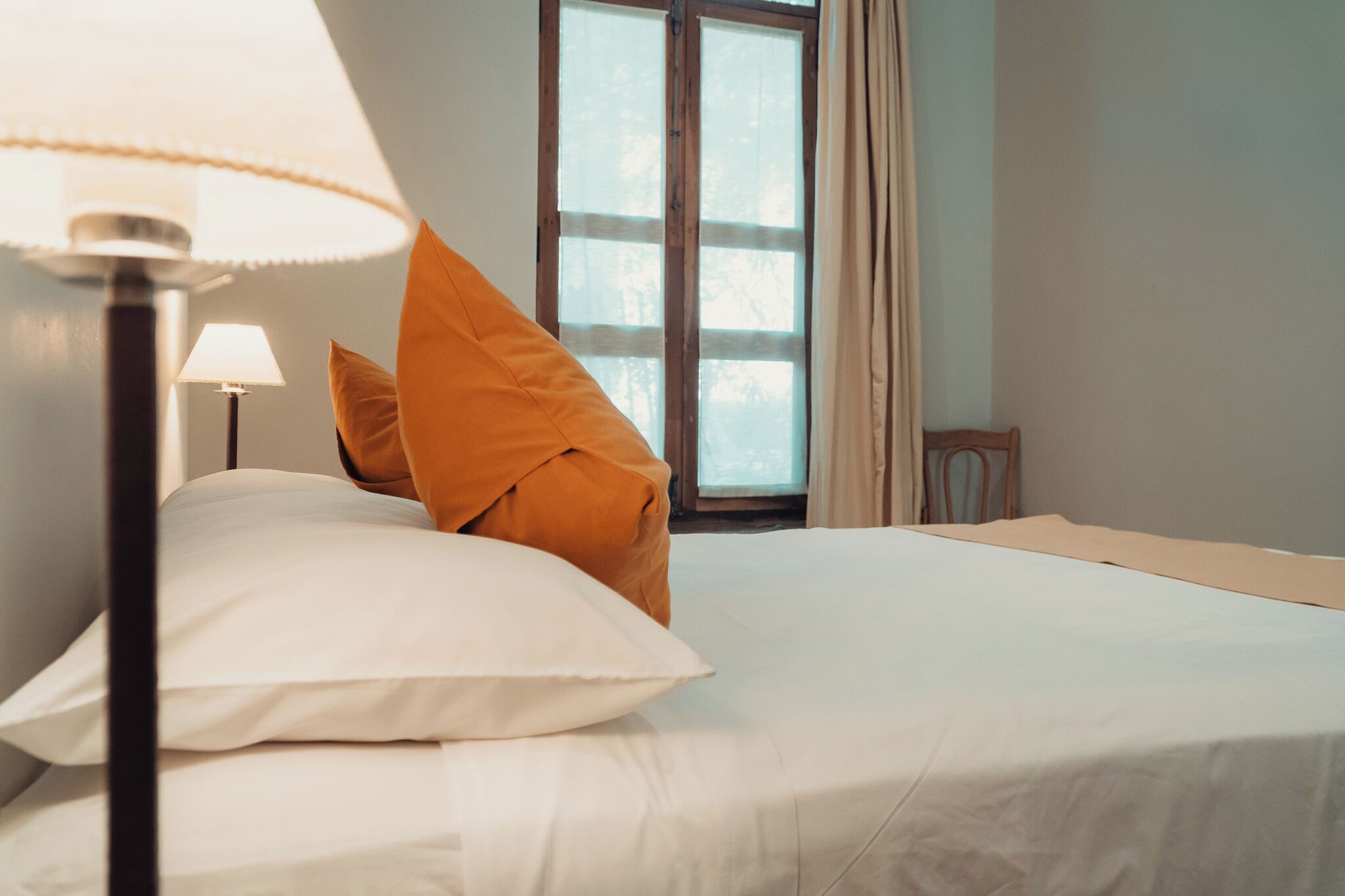 A neatly made bed with white and orange pillows, a lamp, and a window with curtains.