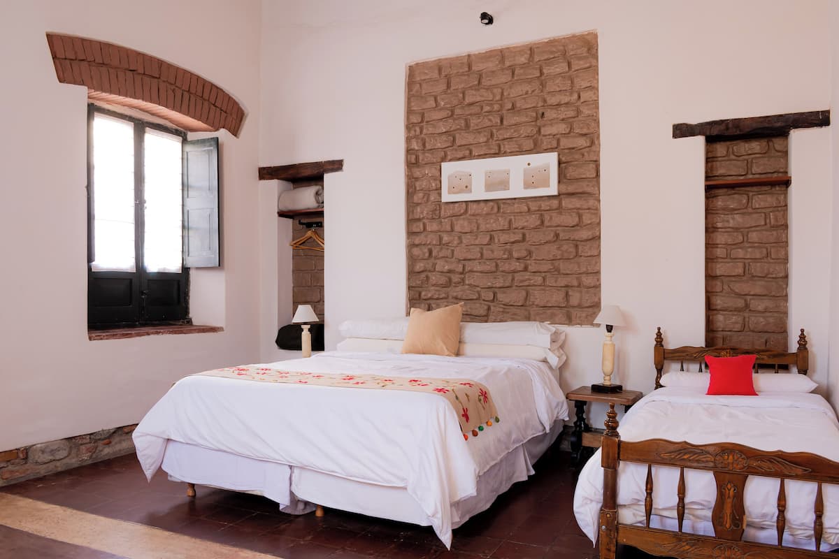 A bedroom with a large bed, a smaller bed, a brick wall, a window, and a wooden headboard.