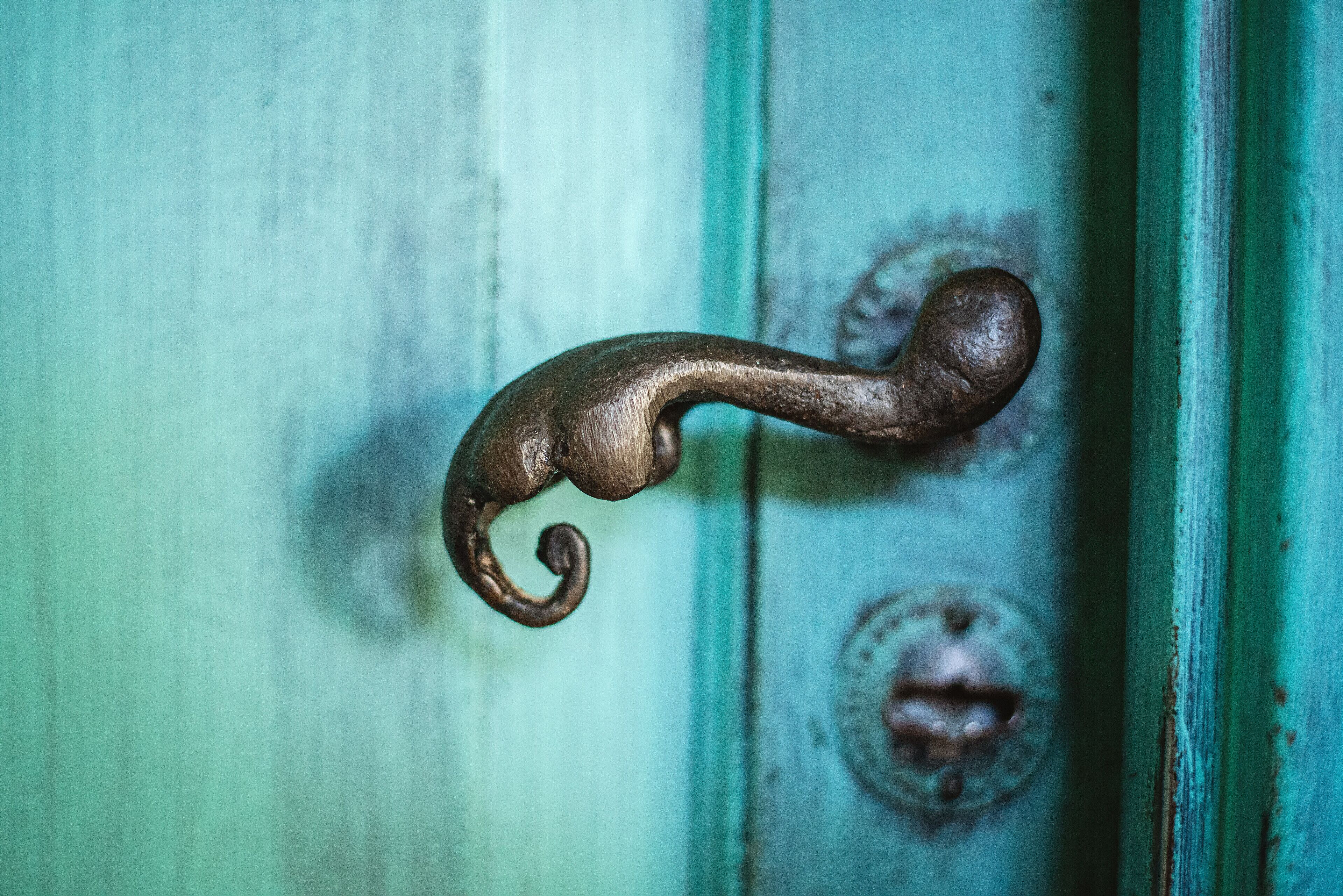 Gros plan d’une poignée de porte rustique et ancienne, au design incurvé, placée sur une porte peinte en bleu sarcelle, dont on peut voir les marques d’usure, et dotée d’un mécanisme de verrouillage rond et circulaire.