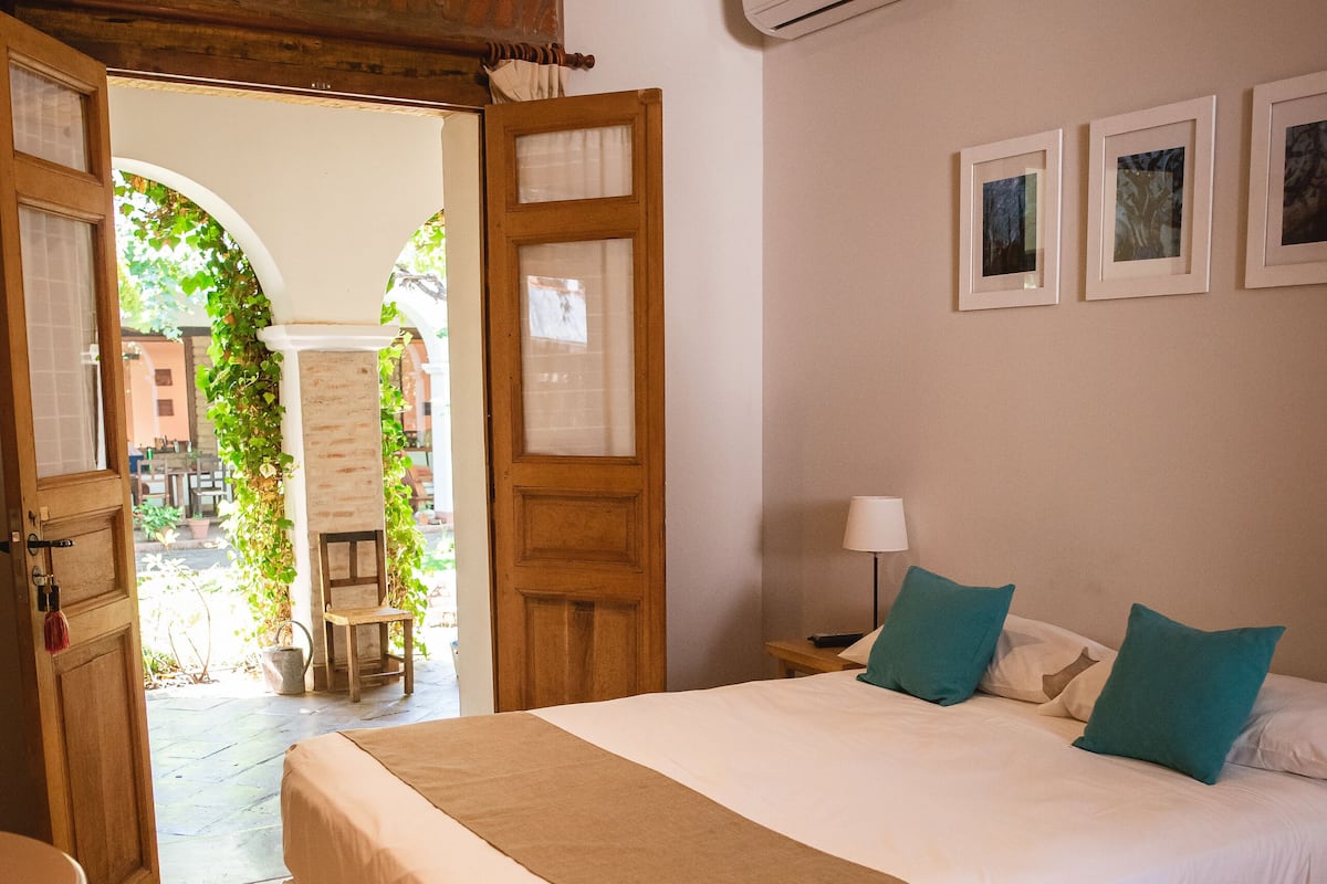 A hotel room with a bed, a nightstand, a lamp, and a view of the outside.