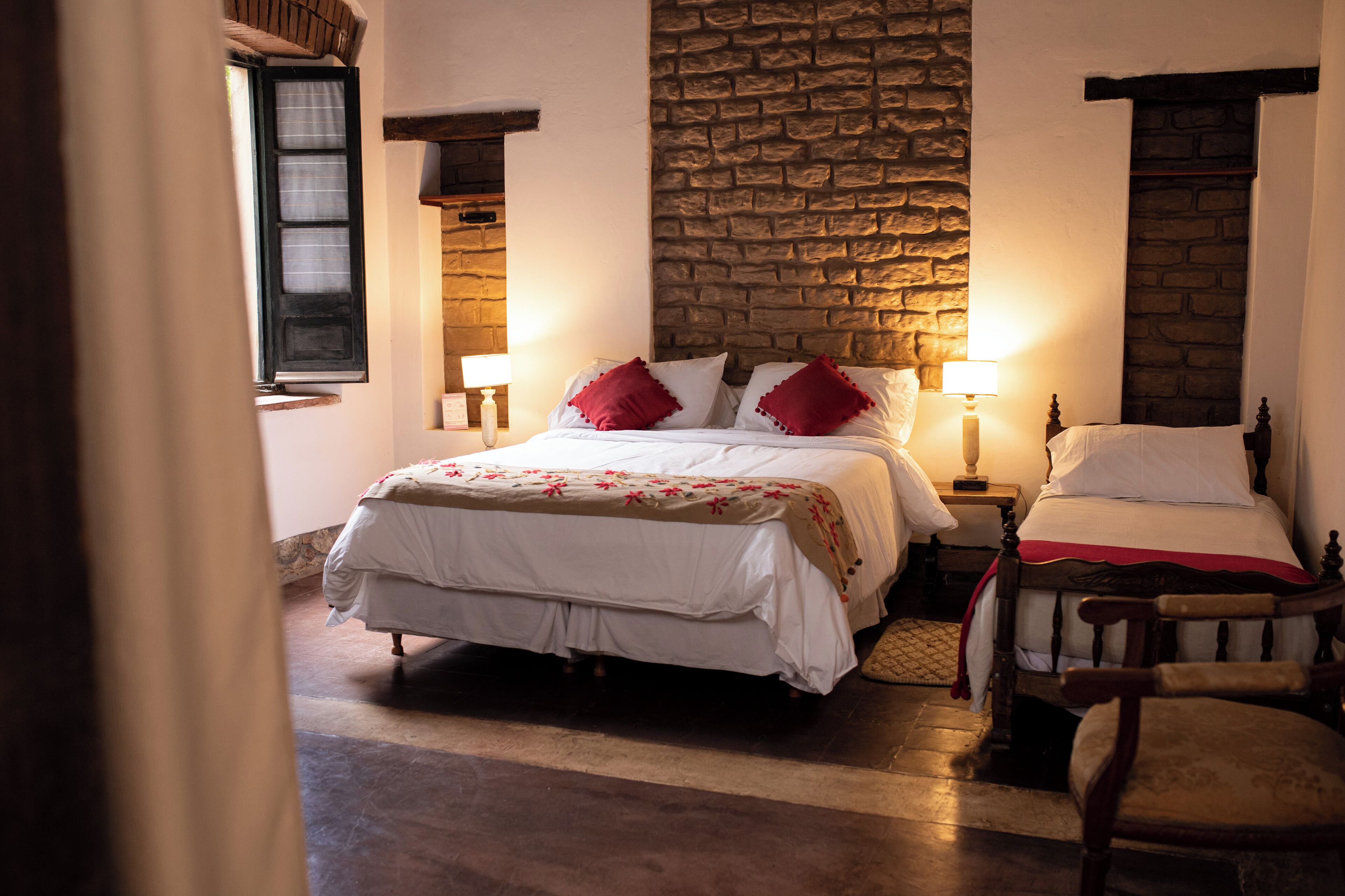 A bedroom with a stone wall, a bed with white bedding and red pillows, a wooden chair, and a nightstand with a lamp.
