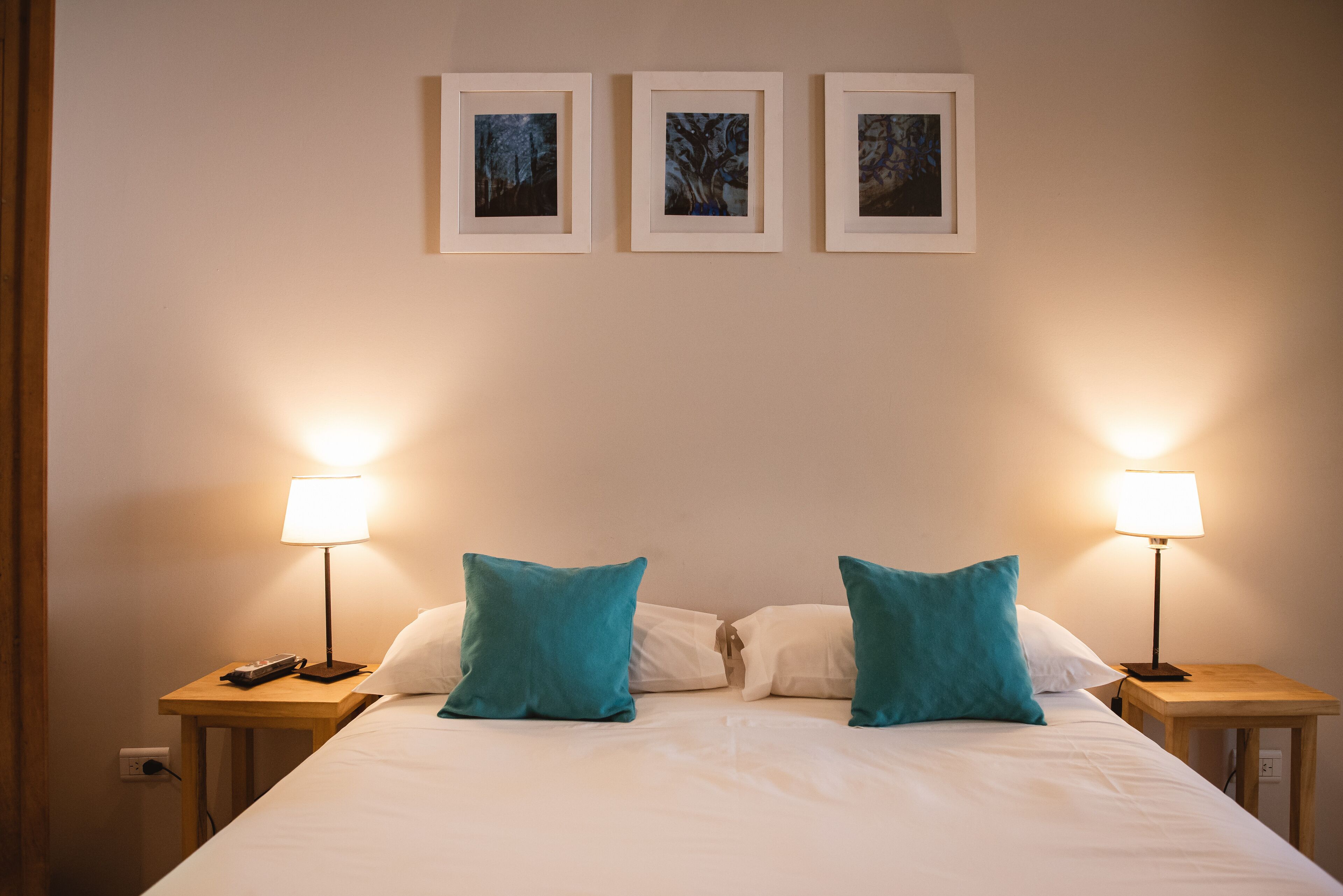 A neatly made bed with two teal pillows, a white comforter, and two bedside lamps on wooden nightstands. There are three framed pictures on the wall above the bed.