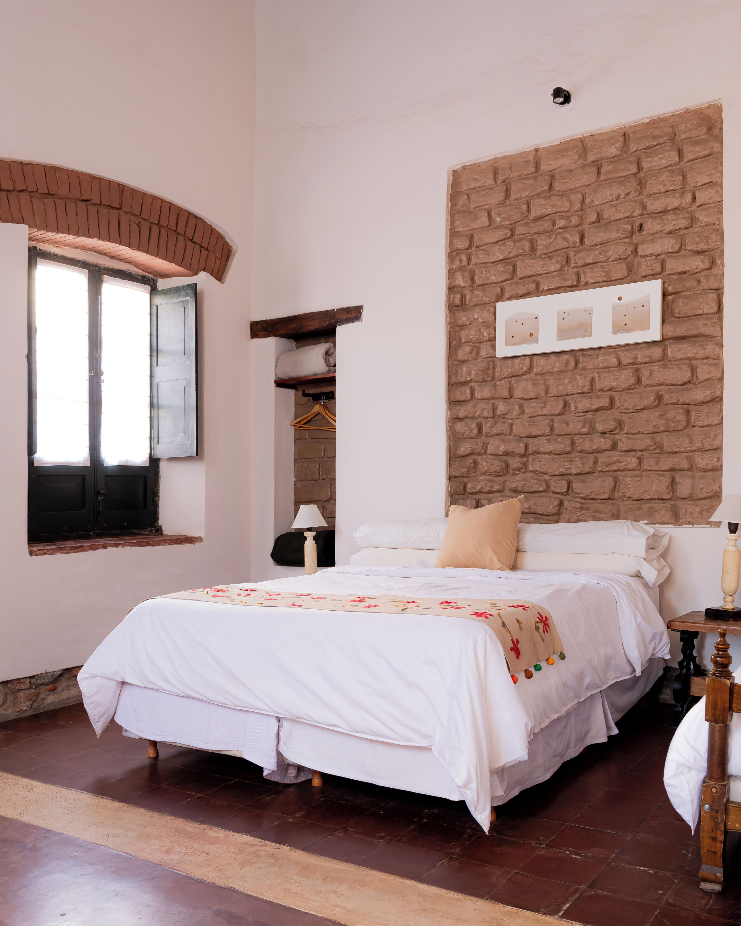 A bedroom with a brick wall, a bed with white bedding, a window with open shutters, and a wooden nightstand with a lamp.