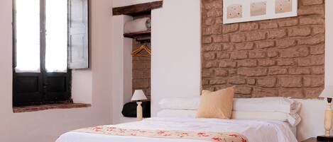 A bedroom with a brick wall, a bed with white bedding, a window with open shutters, and a wooden nightstand with a lamp.