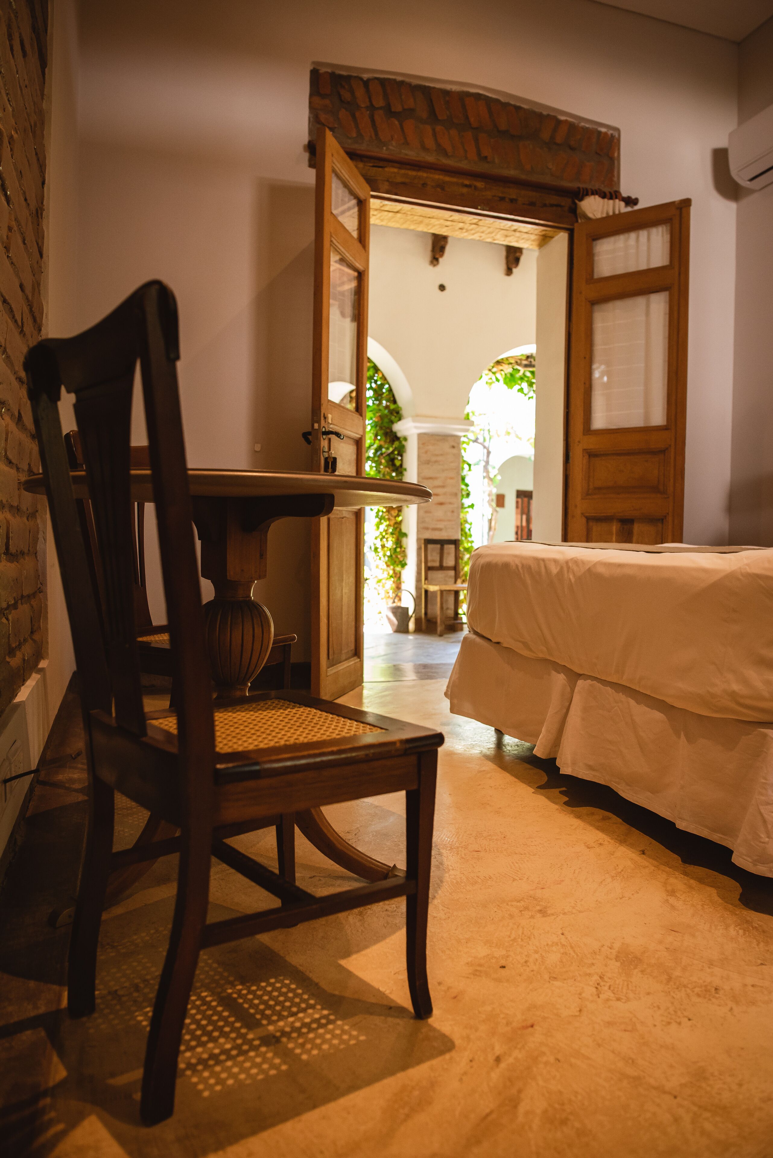 A room with a bed, a wooden chair, a table, and a doorway leading to another room.
