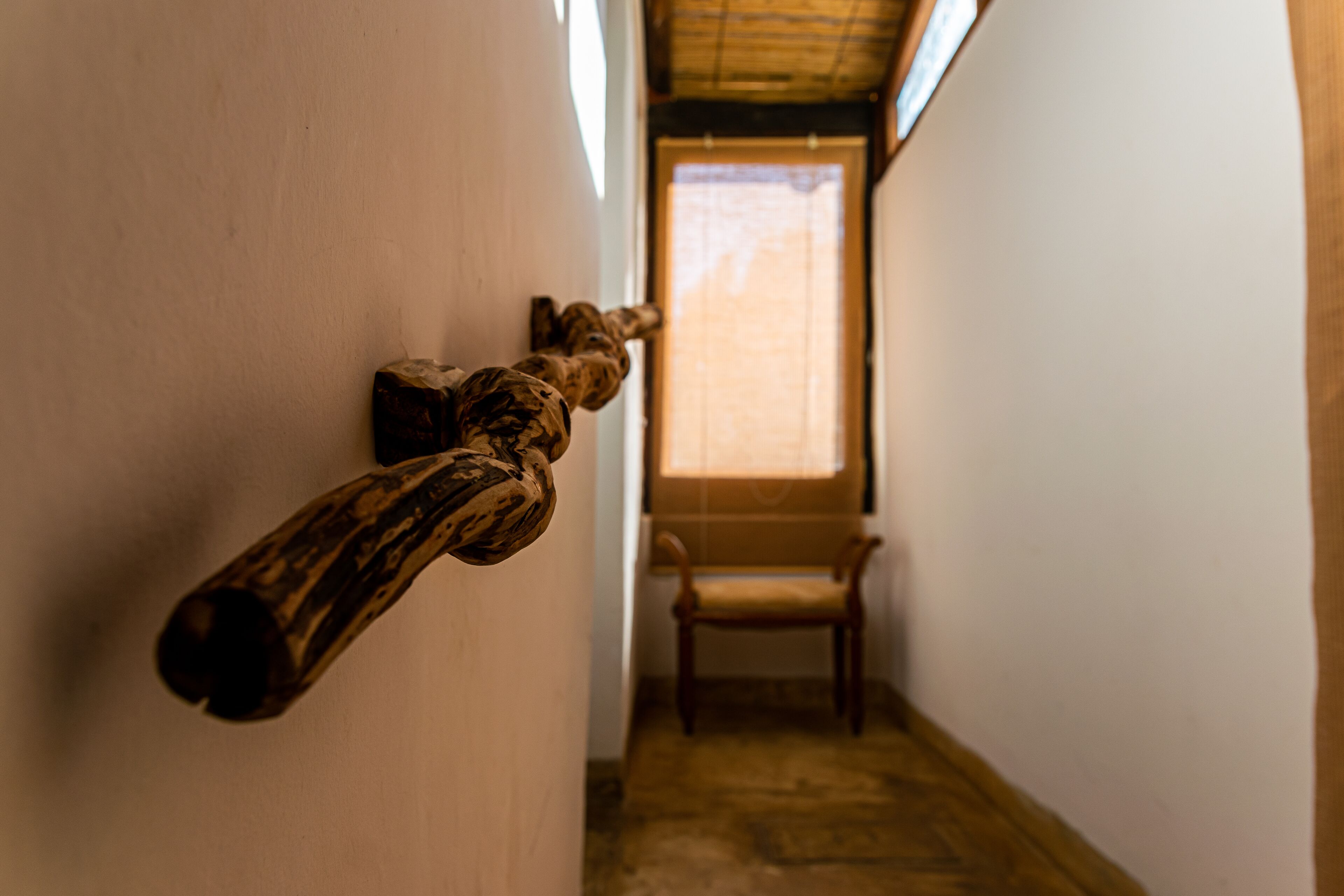 Un pasillo estrecho con una escultura de madera en la pared, una silla de madera y una ventana con luz natural.