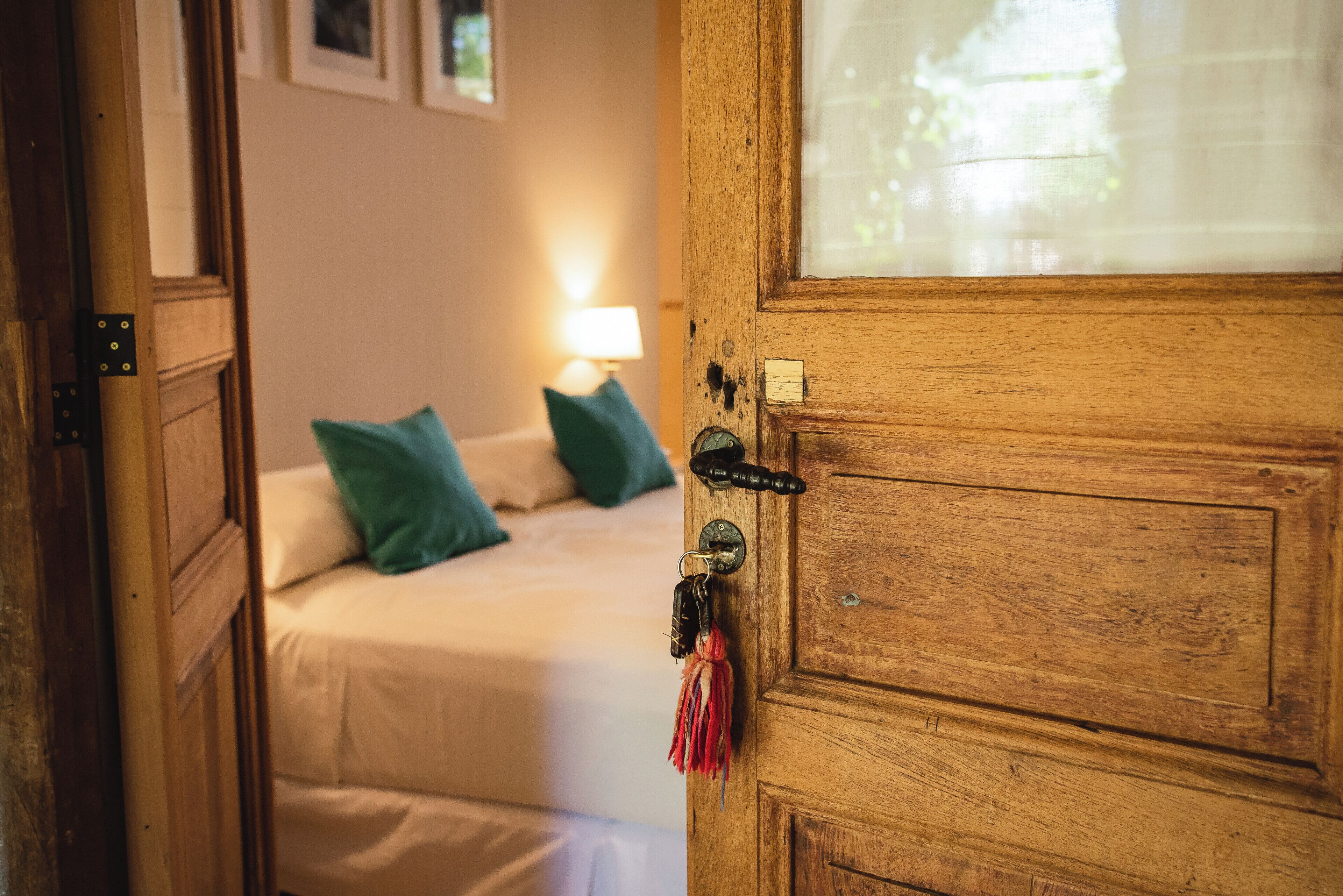 Een houten deur met een sleutelgat en sleutelhanger, die leidt naar een slaapkamer met een bed, kussens en een nachtkastje met een lamp.