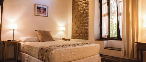 A bedroom with a double bed, two bedside tables with lamps, a window with curtains, a brick wall, and an air conditioning unit.