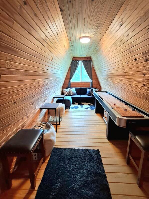 A cozy attic room with wooden walls, a loft bed, a sofa, a table, and a foosball table.
