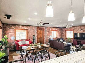 A living room with a brick wall, a red sofa, a brown sofa, a dining table with chairs, a ceiling fan, and a TV.