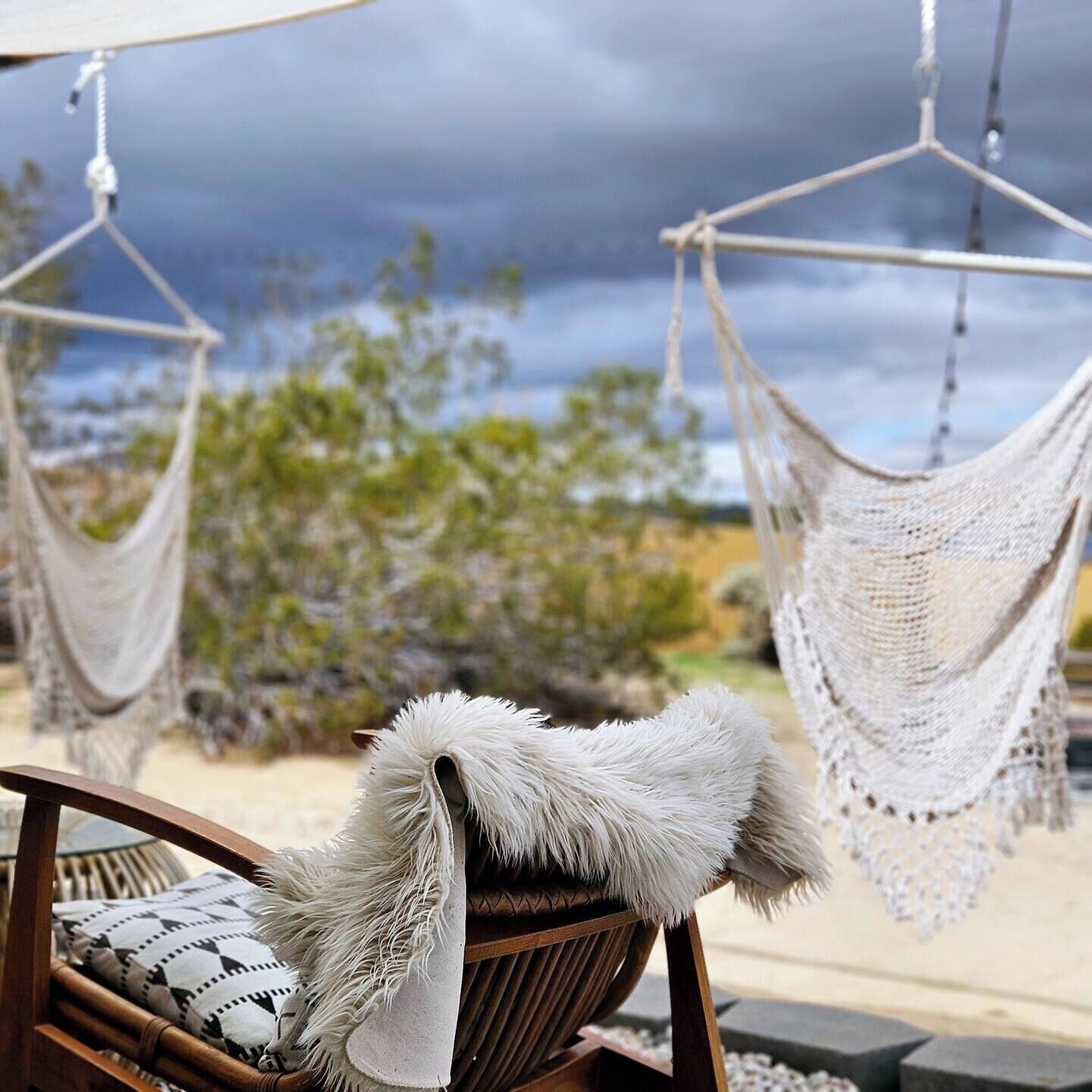 A hammock chair with a white furry throw, a woven chair, and a view of a desert landscape with a cloudy sky.