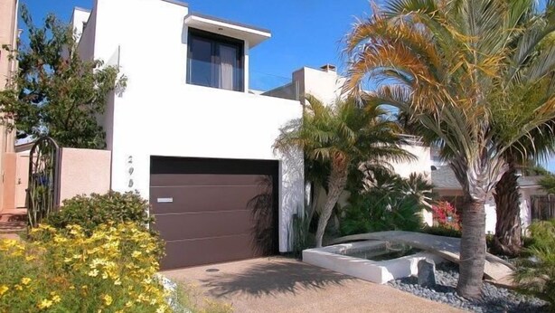 Een modern huis met twee verdiepingen, een garage, omringd door palmbomen en diverse planten.