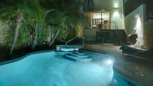 Een zwembad met een jacuzzi, omringd door palmbomen, en een modern huis met een zichtbare keuken en trap.