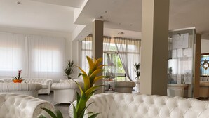 A spacious living room with a large white sectional sofa, a glass coffee table, and several potted plants. - Resort Hotel Tamerici (Marciana Marina)