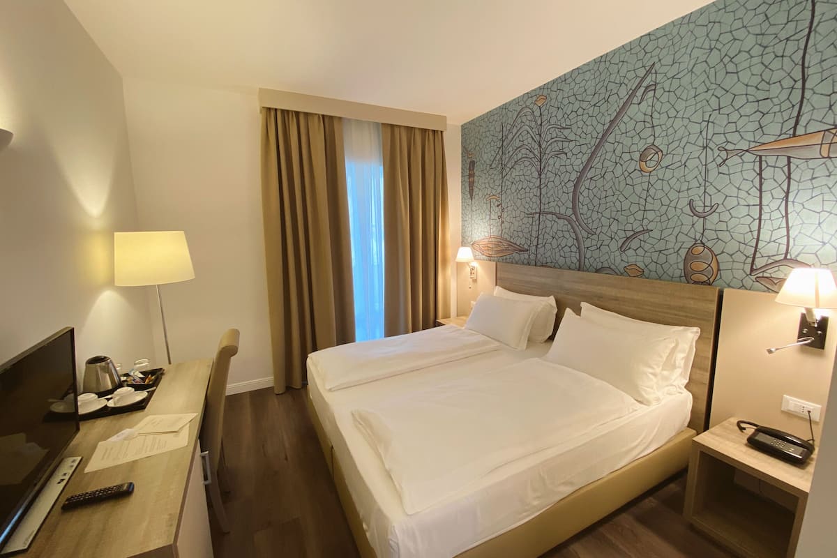 A hotel room with a large bed, a desk with a lamp, a telephone, and a wall with a cracked pattern design.