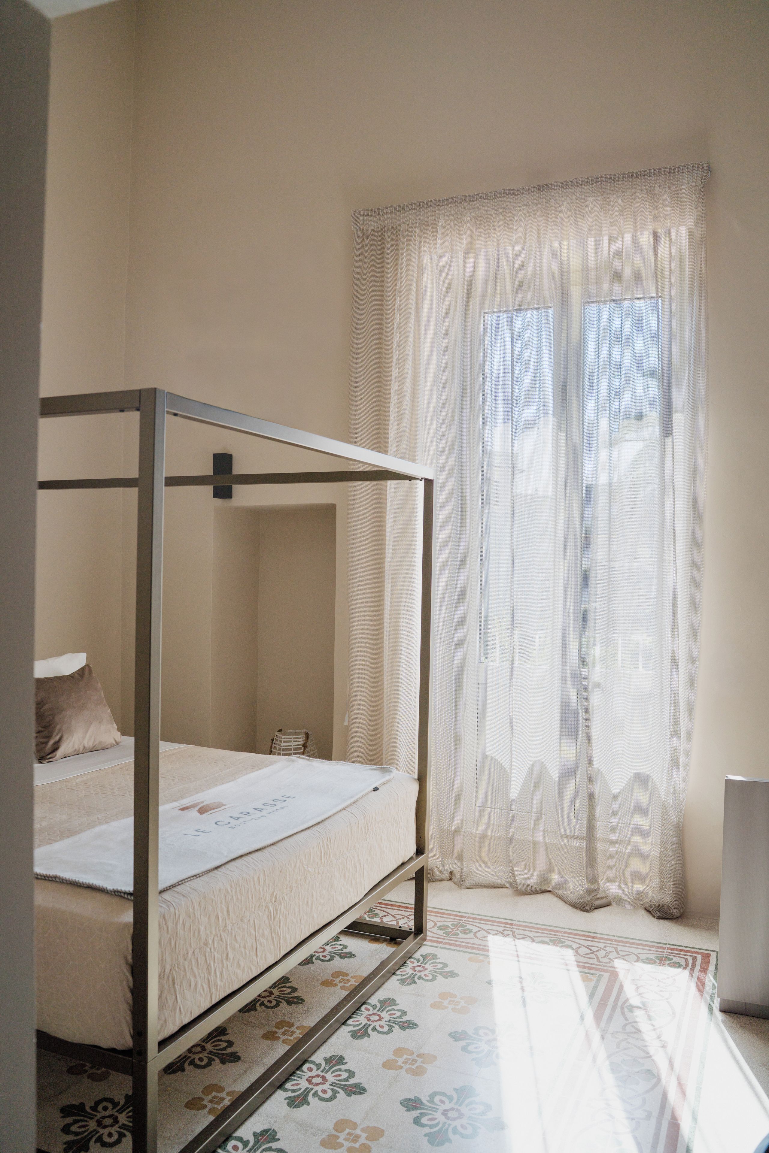 A single bed with a metal frame in a room with a window covered by sheer curtains.