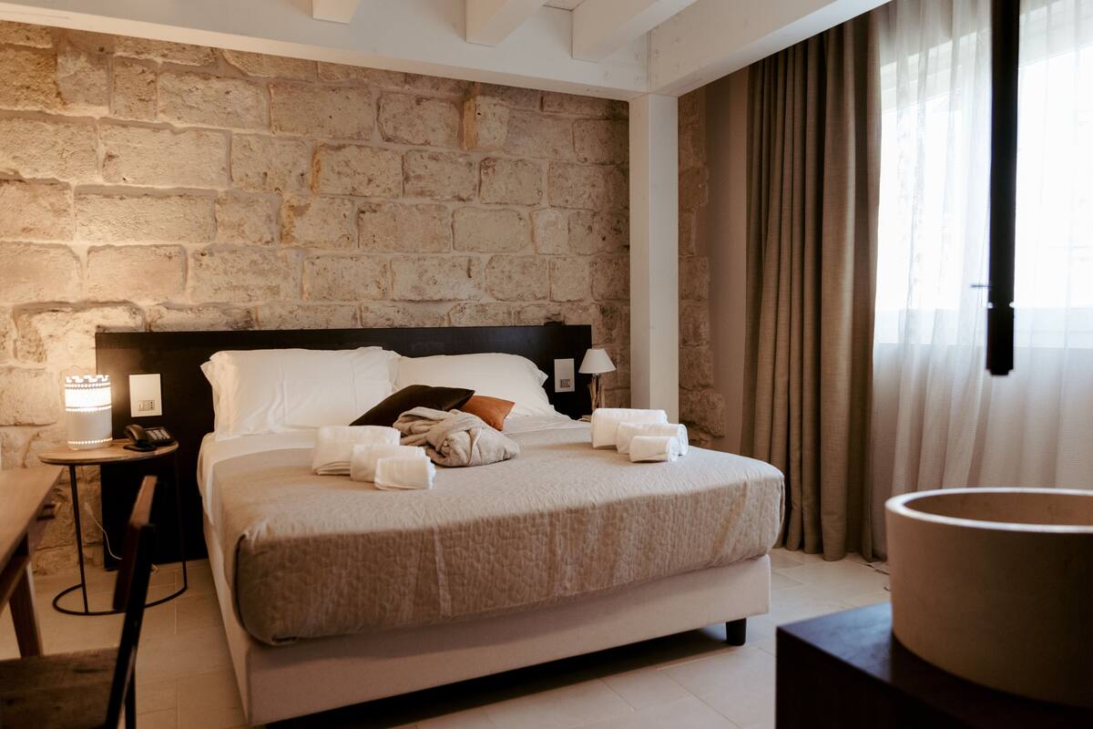 Une chambre d’hôtel avec un mur en pierre, un lit avec du linge de maison blanc, une table de chevet avec une lampe et une grande fenêtre avec des rideaux.