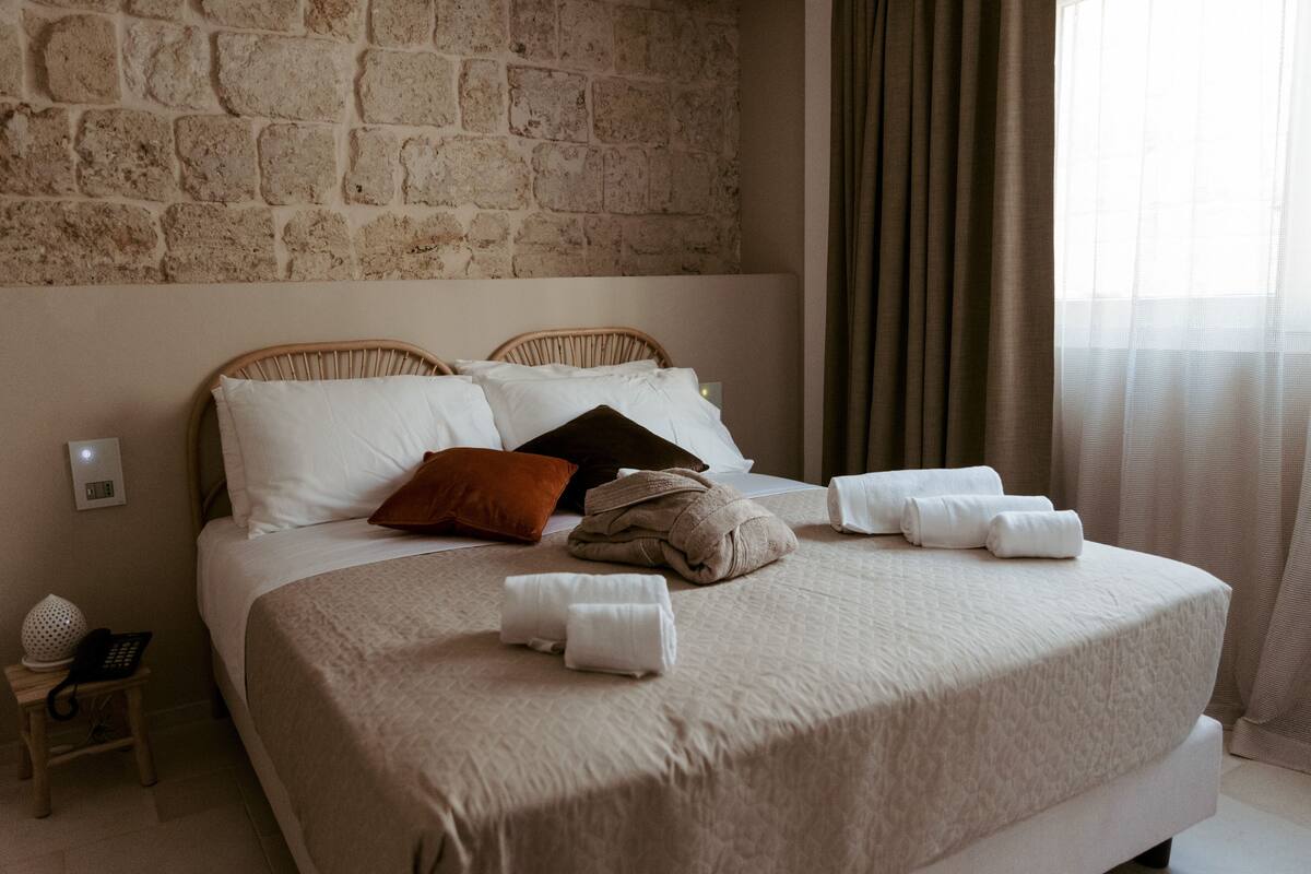 Un lit bien fait, avec une literie blanche et marron, une petite table de chevet avec une lampe, et un mur en pierre derrière le lit.