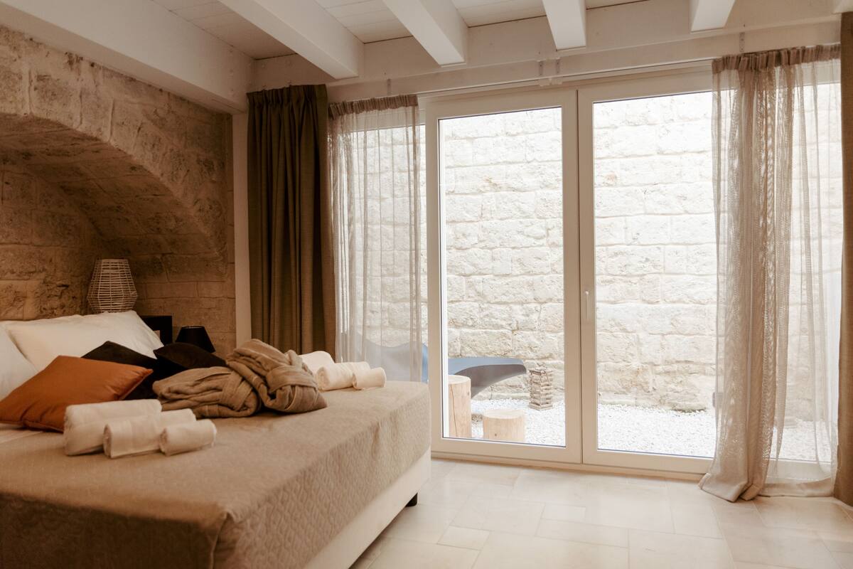 Une chambre à coucher avec un lit, des oreillers, une couverture, une lampe et une vue sur l’extérieur grâce à une grande fenêtre.
