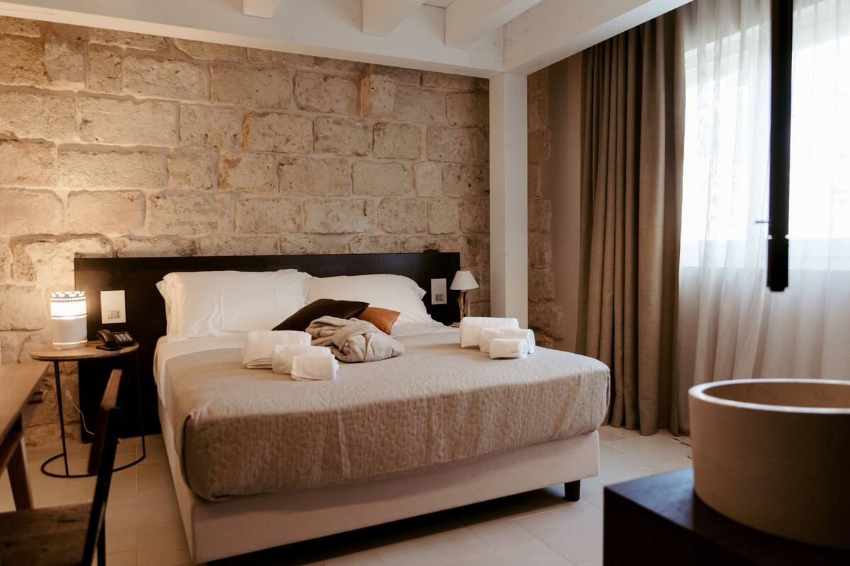 Une chambre d’hôtel avec un mur en pierre, un lit recouvert de draps blancs, une table de chevet avec une lampe, et une vue sur l’extérieur par une fenêtre ornée de rideaux.