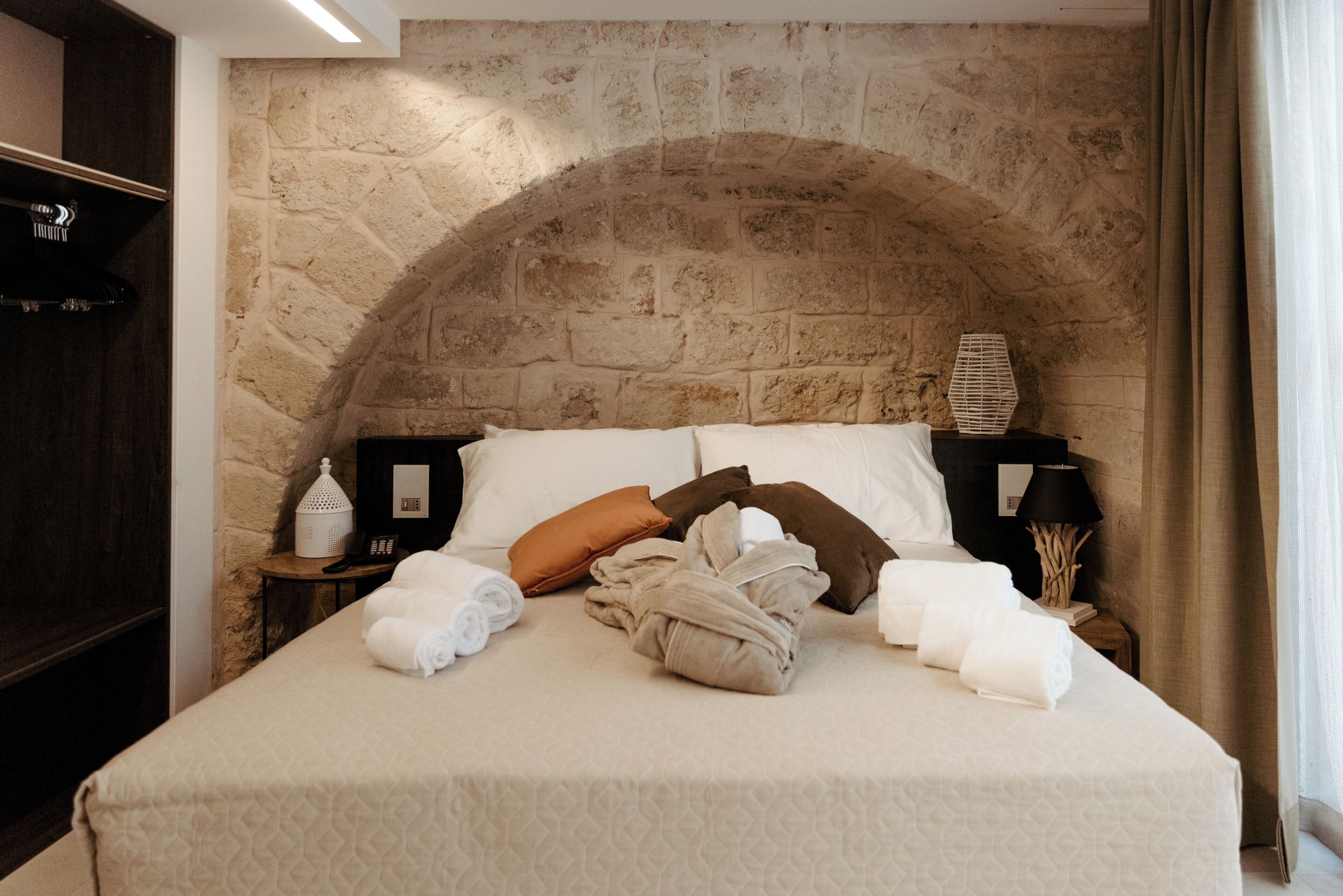 A hotel room with a stone wall, a bed with white linens, a nightstand with a lamp, and a small table with a decorative item.