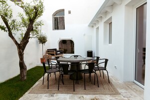 Un patio con una mesa redonda y sillas, un árbol pequeño, y un edificio blanco con una ventana y una puerta.