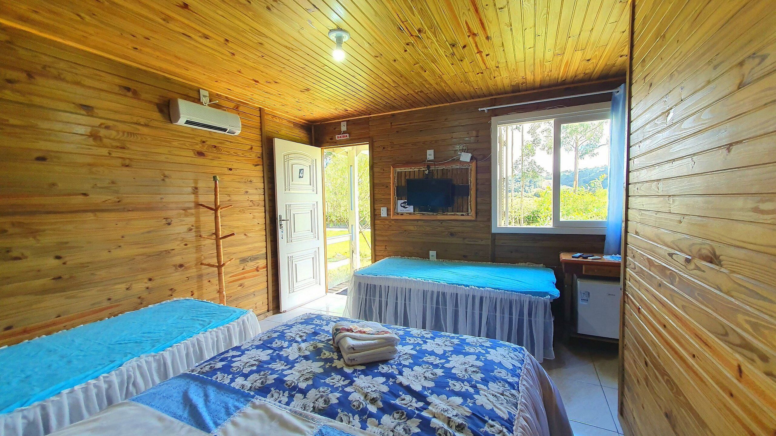 A wooden cabin room with two beds, a TV, and a window with a view of greenery.