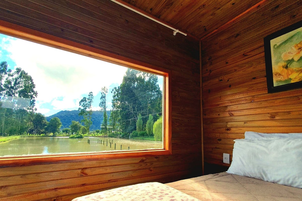 Una habitación de cabaña de madera con una cama, una ventana con vista a un lago y un cuadro en la pared.