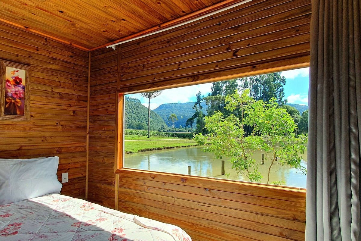 Una habitación de cabaña de madera con una cama, un ventanal con vistas a un río y árboles, y dos cuadros enmarcados en la pared.