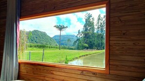 A room with a large window overlooking a green landscape with a water body and trees. - Pousada Mato Verde - Urubici - SC (Urubici)