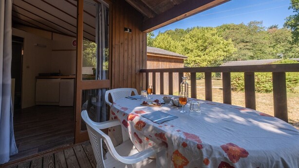 Une terrasse en bois avec une table dressée pour deux personnes, un petit coin cuisine et une vue sur les arbres et les collines.