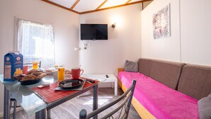 Un salon chaleureux avec un canapé, un téléviseur fixé au mur et une table en verre servant de coin repas pour le petit-déjeuner.