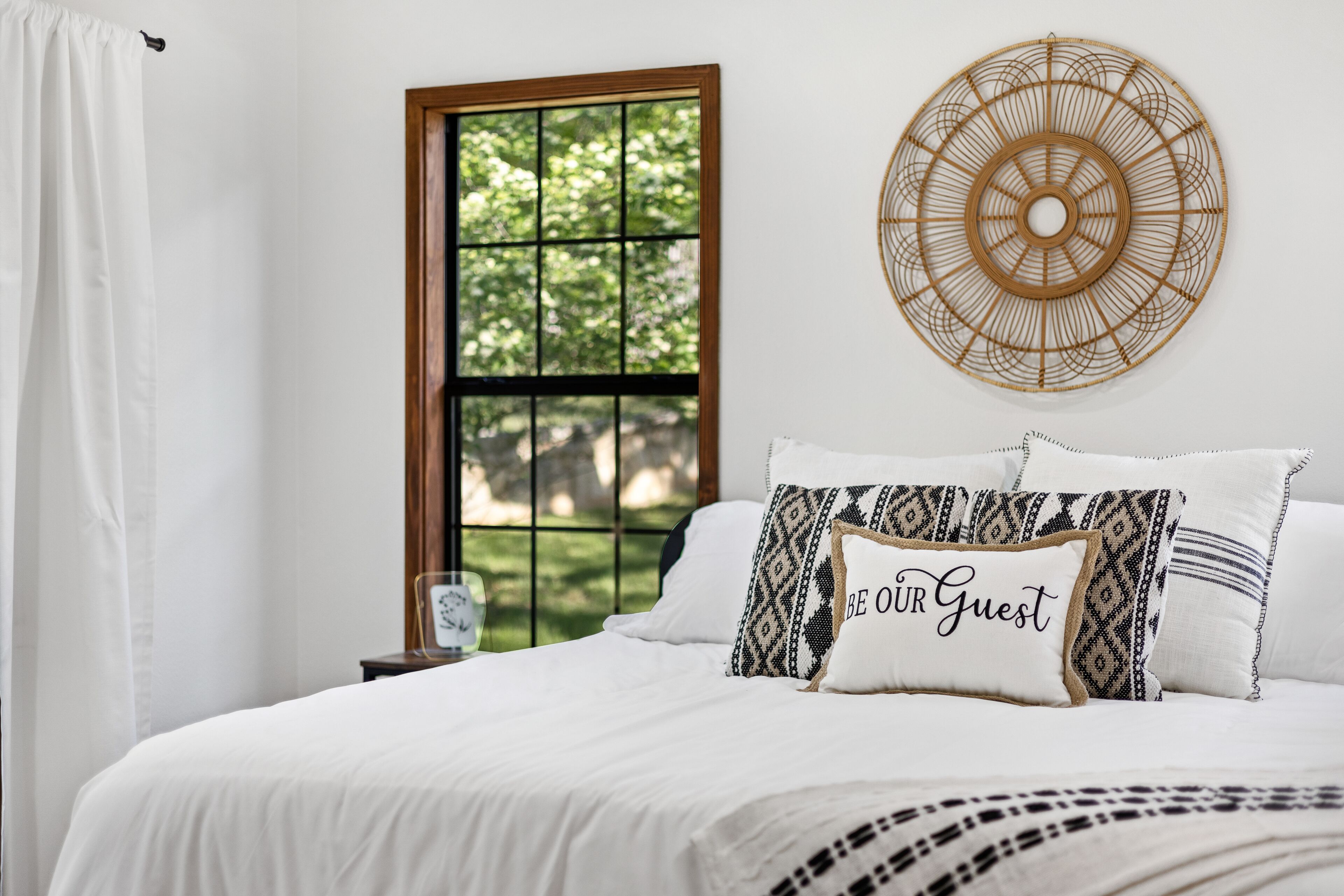 A neatly made bed with patterned pillows, a wall-mounted decorative item, and a window with a view of greenery.
