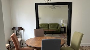 A dining area with a round wooden table and four chairs, a chandelier, and a view into a living room with a green sofa and a ceiling fan.