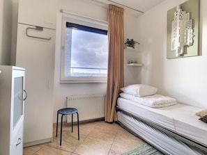 Habitación pequeña y funcional con cama, radiador, ventana con cortinas, un taburete y estante en la pared.